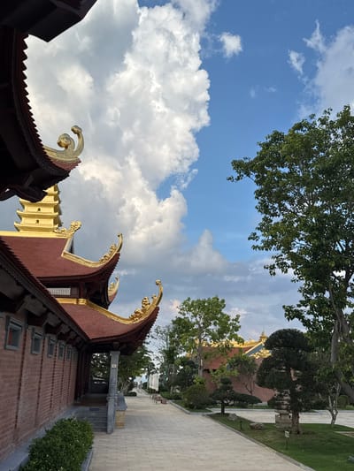 The image shows a view of a temple in Diên Khánh, Vietnam. The temple features ornate architecture with red tiled roofs and golden decorative elements on the eaves. A golden pagoda-like structure is visible in the background. The foreground shows a paved walkway lined with manicured trees and grass. The sky is a mix of blue with dramatic white clouds. The overall scene conveys a sense of tranquility and cultural richness.