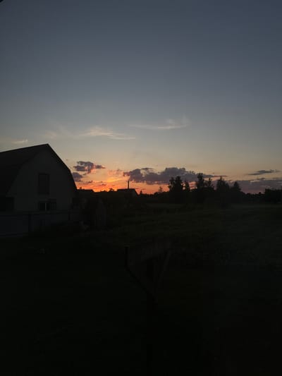 The image depicts a rural landscape at dusk in the village of Chemerne, Ukraine. In the foreground, a dark, silhouetted structure, possibly a fence or a low wall, occupies the lower part of the frame. To the left, a large, dark building with a pitched roof is visible, casting a shadow. The midground shows a dark expanse of vegetation, likely fields or a garden. In the distance, a line of trees creates a dark silhouette against the horizon. Above the trees, the sky transitions from a muted orange and pink near the horizon, where the sun has set, to a gradient of blues and grays towards the top of the image. Scattered clouds are present, some tinged with the colors of the sunset. Several indistinct structures, possibly other buildings or utility poles, are visible among the trees. The overall impression is one of twilight in a quiet, rural setting.