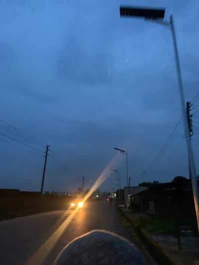 The image depicts a rural road scene at dusk or dawn in Barikin Jirga Summa, Nigeria. The sky is a deep, cloudy blue. A paved road runs through the scene, with a car's headlights illuminating the path and creating bright streaks of light. On the right side of the road, there are several buildings, appearing to be simple structures with dark roofing, and vegetation. Utility poles with multiple wires are visible along the road. A modern street lamp with a rectangular fixture is prominent on the right side of the image, and other street lamps are spaced along the road in the distance. The left side of the road features a dark, low-lying structure and more utility poles. A fuzzy, rounded object, possibly the top of a person's head or shoulder, is in the foreground, obscuring part of the road.