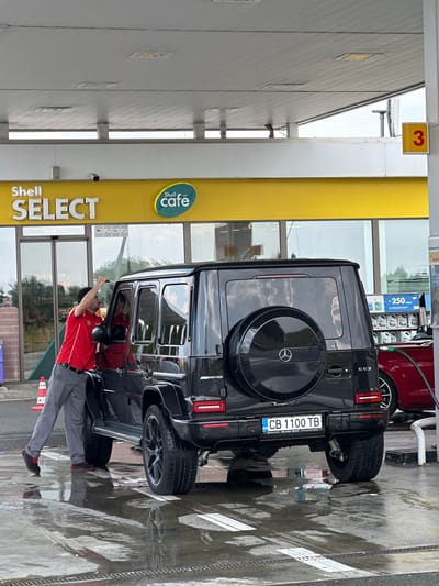 A male attendant in a red Shell uniform with gray pants is wiping the passenger side window of a black Mercedes-Benz G-Class SUV. The vehicle has a spare tire mounted on the rear and a license plate reading "CB 1100 TB". The scene appears to be a Shell gas station, indicated by the "Shell SELECT" and "Shell cafe" signage visible in the background. The ground is wet, suggesting recent rain or cleaning. A red sports car is partially visible to the right of the SUV, and a gas pump hose is connected to it. The overall environment is an outdoor covered area at a service station. The location is identified as Drazhevo, Bulgaria.