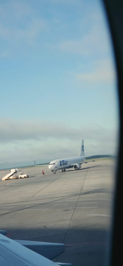 An airplane with "UTair" branding is parked on an airport tarmac under a hazy blue sky with scattered clouds. The aircraft is a commercial jetliner, likely a Boeing 737, with its engines visible. To the left of the plane, a mobile airstair unit is positioned, connected to a small utility vehicle. A single individual, wearing an orange safety vest and dark pants, stands between the airstair and the airplane, appearing to be ground crew. The tarmac is a grey concrete surface with painted white lines. In the foreground, a portion of another aircraft's wing and fuselage is visible, suggesting the photo was taken from inside an airplane. The background shows a distant tree line and a hazy horizon. The scene depicts a typical airport operational environment during daylight hours, possibly at Istanbul Airport, which serves Arnavutköy, Turkey.