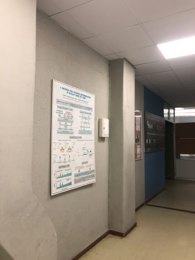 The image shows a hallway with a concrete wall on the left, a large poster on the wall, a large pillar on the right, and a blue wall with a poster on the right. The poster on the left has the title, "A METHOD FOR URANIUM DETERMINATION IN HUMAN CRIME BY ICP-OES" and displays scientific diagrams. The poster on the right has various circles with different images and captions. The hallway is empty and appears to be in a laboratory or academic setting. The coordinates indicate a location in the city of Agia Paraskevi, Greece.