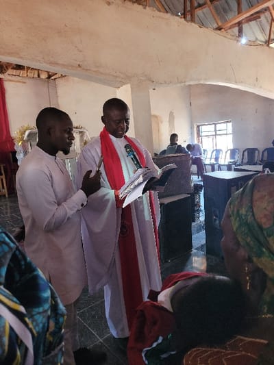 An indoor gathering is depicted in Rinze, Nigeria, within a building featuring light-colored, textured walls, a dark tiled floor, and an exposed wooden rafter ceiling. Centrally positioned, an older male, dressed in a white alb and a red stole, reads from an open book while speaking into a handheld microphone. To his left, a younger male, wearing a light-colored traditional garment, stands observing, holding a mobile phone in his right hand. In the background, several individuals are seated on white plastic chairs. A multi-paned window is visible on the right wall, and a black speaker box is situated near the foreground figures. A small white structure with golden embellishments is partially visible on the far left.