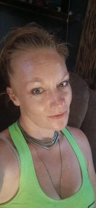 This image captures a single adult Caucasian female, mid-frame, in an indoor setting. She appears to be looking directly at the camera with a slight smile. Her hair is light-colored and pulled back, with some loose strands visible on her forehead. She has light-colored eyes and minor facial freckles. The subject is wearing a bright lime green tank top or sports bra, along with multiple silver-toned necklaces: one is a multi-band choker style, and at least one other is a long, delicate chain. The background is largely dark and out of focus, revealing what appears to be a dark brown upholstered surface, likely a couch or chair, to the subject's right. Vague, darker shapes in the upper left suggest other furniture or objects. A thin, faint vertical line is present in the upper right background. The specific location within the city of Santa Fe, United States, cannot be determined from the visual evidence presented in the image.