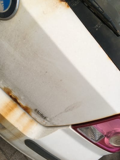 The image shows a close-up view of the front of a white car, likely parked outdoors in Baranavichy, Belarus. The car's hood is visible, along with a portion of the windshield and a taillight. There is significant rust and dirt accumulated on the hood, particularly along the edge where it meets the front of the car. The white paint is discolored and appears faded. A shadow from the windshield Wiper blade is cast diagonally across the hood. A chrome emblem with blue detailing, possibly a car manufacturer's logo, is partially visible in the upper left corner. The text "1.5i" is embossed on the hood near the rust. The taillight, visible in the lower right, is tinted pink and has a clear section for the reverse light. The overall impression is that of an older, somewhat neglected vehicle. The lighting suggests it is daytime, and the presence of dirt and rust might indicate an outdoor location exposed to the elements.