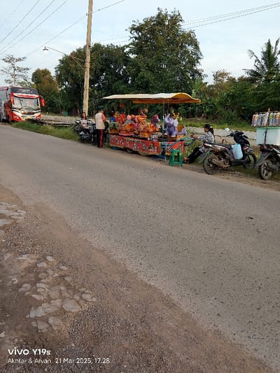 A red bus is parked on the side of a paved road. Several people are standing or sitting near a brightly colored children's ride that is in the shape of a carousel. The ride is parked on the shoulder of the road in front of the bus. The ride is facing the road, and the bus is facing the opposite direction. There are two motorbikes parked near the ride. The scene appears to be in a rural area, with trees on both sides of the road and green grass along the roadside. The photo was taken in Kecamatan Haurgeulis, Indonesia, located at latitude -6.421 and longitude 107.996.