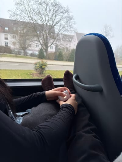 From an elevated perspective within a bus, a person rests their feet, clad in brown socks, against the window. They hold a white, possibly electronic device, with both hands. The bus seat above is blue and grey. Outside the window, trees and buildings are visible against a foggy sky. The person wears a black long-sleeved shirt.