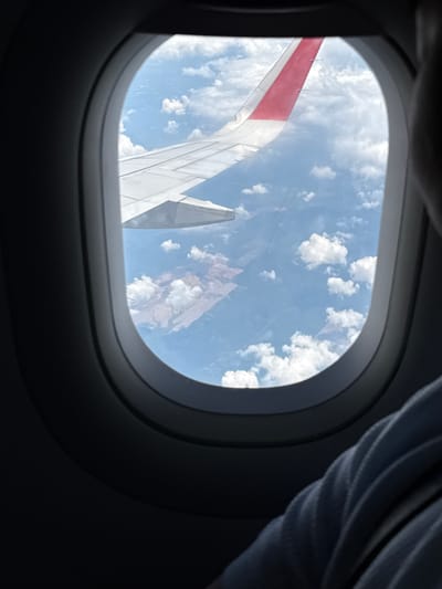 The image shows a view from an airplane window, featuring the aircraft's wing against a backdrop of blue sky and scattered clouds. The window is oval shaped and trimmed in dark gray. Part of a seat or clothing is visible at the top right of the image. The red and white wing with mechanical parts confirms it's an airplane wing. No specific location details relating to Sepang, Malaysia are discernible. No people or interactions are visible. The environment consists of the atmospheric sky and clouds.