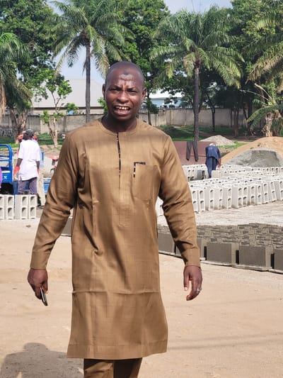 A man in a long-sleeved, brown, plaid-patterned shirt is standing in Kwamba, Nigeria, holding a smartphone. Palm trees are visible in the background. A pile of gray concrete blocks is to the side, near a concrete walkway. Another person, dressed in blue, is visible walking on the concrete blocks. A person wearing a white shirt and a face mask is seen in the background, near a blue cart filled with gray concrete blocks. The ground is light brown.