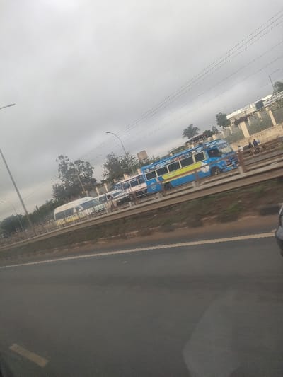 The image depicts an outdoor scene in Biashara ward, Kenya, under a predominantly overcast sky. The immediate foreground consists of a dark asphalt road surface with a faded white or yellow lane marking on its left side. A partial view of a dark vehicle element is visible in the bottom right corner of the frame. A brown median strip with sparse green vegetation and a metal guardrail separates the foreground road from a parallel elevated road or service lane. This lane features a line of vehicles, primarily minibuses (matatus). The most prominent vehicle is a blue and yellow minibus extensively decorated with graphics, positioned centrally. Behind it, a white minibus with a yellow stripe is visible, along with other partially obscured vehicles. Approximately 3-5 individuals are standing near the right side of the blue minibus, adjacent to a metal fence. Utility poles and streetlights are present along the road. In the background, behind the fence, a light-colored building complex is visible, featuring a sign with partially legible text, appearing to read "LIQUOR SO" on its visible portion, mounted on the roof. Additional trees and a water tower are visible further in the background.