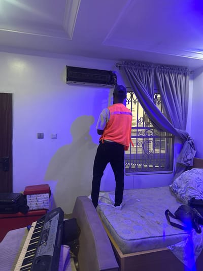 The image depicts the interior of a room in Guzape, Nigeria, featuring a man in a safety vest inspecting or working on an air conditioning unit mounted high on the wall. The room is illuminated with a strong blue light. The man is wearing dark clothing, a cap, and a bright orange safety vest. To his right is a bed with bedding and a bag. A window with ornate metal bars is partially covered by grey curtains. Below the air conditioner, there are electrical outlets and a dark wooden door. A keyboard is in the right corner of the room. Suitcases and other objects are scattered nearby.