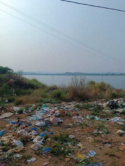 The scene depicts a significant amount of mixed waste scattered across bare earth in Kothacheruvu, India. The debris includes plastic bags, packaging materials, paper, and other household waste. In the background, a body of water is visible, with vegetation growing along the edge where the trash is deposited. The background also shows a distant horizon, power lines and hills under a slightly hazy sky. No people or animals are visible. The environment appears neglected, with the accumulation of trash negatively impacting the landscape near the water source.