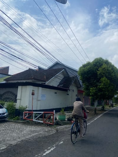 The image shows a residential street in Mojokerto, Indonesia, at the location of latitude: -7.475, longitude: 112.441. A person is riding a bicycle down the street. The person is wearing a brown jacket and a red bag is attached to the front basket of the bike. There are several homes to the left of the street, and a tree to the right. There is a white car parked to the left of the frame. Many power lines stretch across the sky. There are other homes visible in the distance. The sky is cloudy and blue.