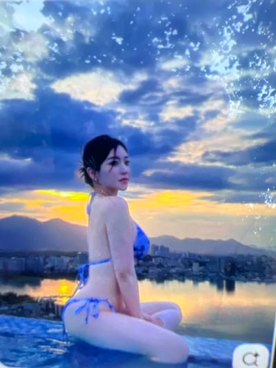 The image shows a young woman in a blue bikini sitting in an infinity pool. She is looking to her right, away from the camera, with her head tilted slightly. Her hair is dark and pulled back in a messy bun. She is wearing dark eye makeup. The background depicts a city skyline with mountains in the distance. The sky is cloudy with hues of orange and yellow from a sunset or sunrise. The water of the infinity pool reflects the sky and the city. There is a circular icon with an arrow in the bottom right corner of the image. The location is not Kara-Dıykan, Kyrgyzstan; this scene appears to be a city with a body of water and mountains, possibly a coastal or lakefront urban area.