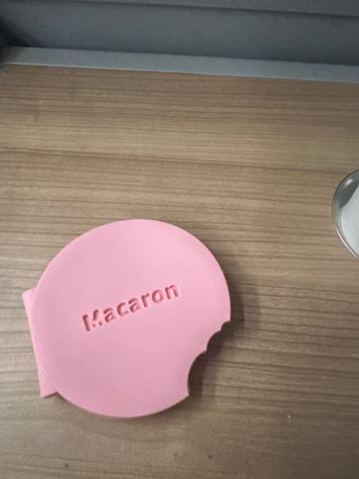 The image shows a pink, round object with the word "Macaron" embossed on its surface. A portion of the object is missing, creating a notched or bitten appearance on the upper right side. The object rests on a light brown wooden surface, likely a table or desk. Part of a gray object is visible on the top and left. The location appears to be Mate Yehuda Regional Council, Israel.