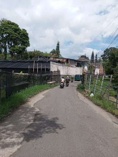 The image depicts a paved, two-lane road in Ariyalai, Sri Lanka, under a partly cloudy sky. The road, showing wear and tear with visible shadows, curves slightly to the right in the distance. Three individuals on motorcycles are traveling away from the viewer, positioned centrally on the road. The lead motorcyclist wears a light-colored top and a white helmet. The two following motorcyclists wear dark clothing and dark helmets. Ahead of the motorcycles, a green van is parked or stopped on the right side of the road.
To the left of the road, a grassy verge transitions into a black mesh fence, likely agricultural shading, supported by vertical wooden poles. Behind this fencing is a long, low structure with a black corrugated roof. Further left, a large, dense green tree dominates the skyline. To the right of the road, a similar grassy verge borders a metal fence structure, beyond which is overgrown vegetation.
In the mid-ground, behind the structures on the left, a light-colored concrete wall is visible. Above and behind this wall, a collection of buildings with varying architectural styles and materials are present, including structures with rusted corrugated metal roofs and white-walled buildings. Power lines are strung across the upper right portion of the scene. Several tall, slender trees, possibly pine, are scattered throughout the background. Distant hills or mountains are faintly visible under the horizon line.