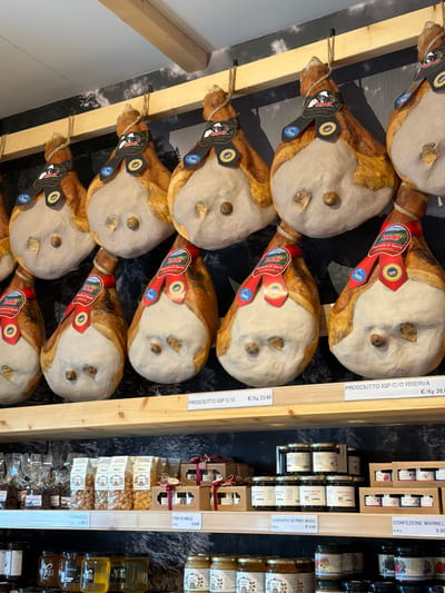 The image depicts a display of cured hams hanging from wooden beams in what appears to be a shop. The hams are large and have a distinctive prosciutto shape, with the fat cap visible and a prominent hoof. Each ham is adorned with a black and red label that reads "Wolf Prosciutto di Sauris". The labels also feature a circular seal with the text "Sauris" and other emblems. The hams are arranged in rows, with some visible on upper and lower shelves. Below the hanging hams, on the lower shelves, are various jars and packaged goods, including what appear to be jams, honey, and possibly dried fruits or nuts. Some of these items also have labels with Italian text. The background features a dark, textured wallpaper with a subtle pattern that might represent a forest or mountainous landscape. Wooden planks run horizontally across the frame, serving as shelves or supports for the display. The overall impression is of a food store or market stall specializing in cured meats and local products. The presence of the "Prosciutto di Sauris" labels strongly suggests the location is in or around Sauris / Zahre, Italy, a town known for its unique cured ham.