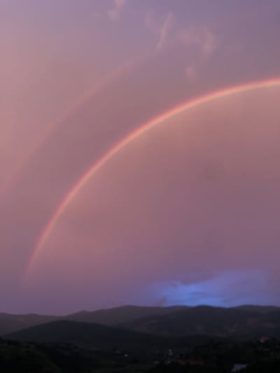 This media file captures a natural phenomenon, likely a double rainbow, set against a backdrop of a hazy sky and a silhouetted mountain range. The sky is dominated by soft, diffused light in shades of purple and pink, suggesting either sunrise or sunset. Wispy clouds are visible, blending with the overall atmospheric haze.
The most prominent subjects are the two arcs of the rainbow, appearing as bands of faint colors. The primary rainbow is more distinct, showing a gradient of reds, oranges, and yellows. A fainter secondary rainbow is visible just outside the primary one, with its colors appearing in reverse order.
On the right side of the image, a dark, imposing mountain range rises steeply. The silhouette is so dark that details are obscured, but some hints of vegetation can be seen along its upper slopes. The mountain is partially shrouded in mist or haze, creating a dramatic contrast with the vibrant colors of the rainbow.
The overall mood is serene and awe-inspiring, evoking a sense of natural beauty and wonder. The time of day is clearly either dawn or dusk due to the quality of light and the presence of a rainbow. The weather appears to be somewhat misty or rainy, which is a prerequisite for rainbow formation.
There is no visible text in the image. The location context of Thôn 4, Vietnam, adds a geographical element, suggesting this natural spectacle is occurring in that region, possibly after a rain shower in a mountainous area. The image focuses entirely on the sky and the landscape, with no human figures or man-made objects visible.