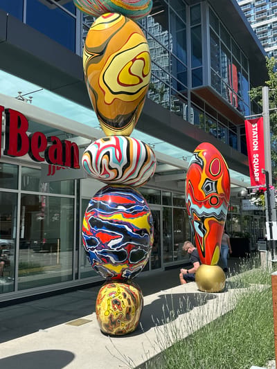 The image depicts an outdoor scene in Burnaby, Canada, specifically at "The Bean" coffee shop located in Station Square. Dominating the foreground are several large, brightly colored, abstract sculptures stacked vertically. These sculptures have a glossy, organic form and are painted with intricate, swirling patterns in a variety of colors including yellow, orange, red, blue, black, white, and pink.

To the left, the red, stylized lettering of "The Bean" is visible, indicating the establishment. Behind the sculptures, the facade of a modern building with large glass windows is visible, reflecting the surrounding environment and other buildings. A red banner with white lettering that reads "STATION SQUARE" is attached to a pole on the right side of the image, further identifying the location.

In the middle ground, a man is seated on a low wall or ledge, looking down at something in his hands, possibly a phone. He is wearing a dark t-shirt and shorts. Another person is partially visible walking behind him. The ground in front of the building is paved, and a patch of green plants, possibly lavender, is growing in the lower right corner. The overall impression is of a public space with art installations in an urban setting.