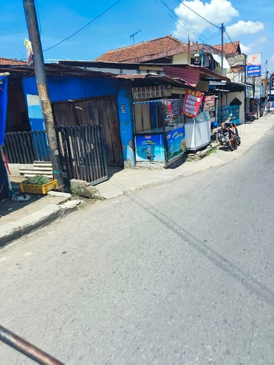 The image shows a street scene in Kecamatan Margahayu, Indonesia, taken from the perspective of a person walking down the street. The image is taken at a slight angle, with a building on the left side of the image and a road in the foreground. The building is painted blue with a brown door and window. The building has a sign that says "MARTABAK BANGKA" in white letters on a red background. There is a motorcycle parked in front of the building. The road is paved and appears to be empty of traffic. The sky is clear and blue with some white clouds. There is a sign on the building across the street that reads "APOTEK". The street is lined with buildings on either side. There are other buildings in the background. There are power lines in the sky.  The image is taken in a residential area and it appears that it is daytime.