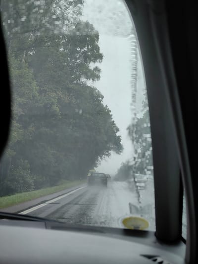 The image shows a view from inside a car looking out the windshield on a rainy day. The windshield is covered in raindrops, distorting the view of the road and the trees lining it. A yellow vehicle is visible in the distance on the wet road, followed by other cars. The scene appears to be on a rural road, possibly in or near the Шчомысліцкі сельскі Савет in Belarus, given the presence of dense green trees and a paved road with white markings. The sky is overcast and appears to be a light grey, consistent with rainy weather. The interior of the car is visible in the foreground, including the dashboard and part of the windshield frame. There are no people visible in the frame.