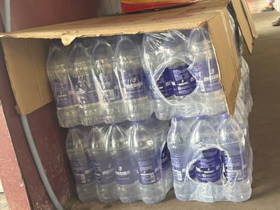 The image depicts a close-up, overhead view of several stacks of bottled water products. The bottles are clear plastic with blue labels that read "MERIBA Life in every drop" and "1 litre". The bottles are packaged in clear plastic wrap and are stacked within an open cardboard box, which is partially supported by a ledge. The background shows a textured reddish-brown surface, possibly a wall, and a grey corrugated pipe running vertically along it. The floor appears to be concrete. The overall scene suggests a storage or distribution point for bottled water. The lighting is somewhat dim, and the focus is on the product.