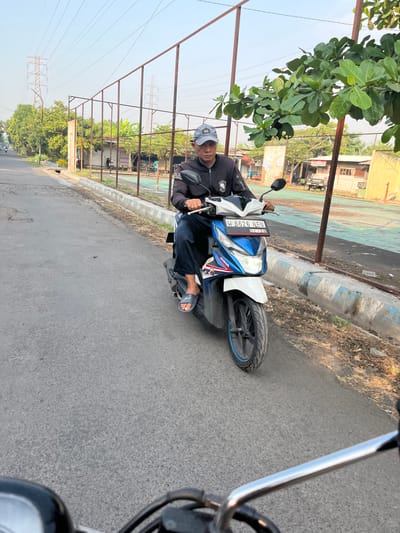 A man wearing a gray baseball cap and a black jacket is riding a blue and white Honda scooter on a paved road in Kedungkendo, Indonesia. He is wearing dark pants and gray sandals. The scooter has a license plate that reads "W 6626 NEU". In the background, there is a chain-link fence surrounding a sports field, with trees and utility poles visible beyond. The sky is clear and the sun is shining. The road appears to be in a semi-urban or suburban area, with some buildings and vegetation lining the side of the road. The perspective of the image suggests it was taken from another vehicle or scooter following behind.