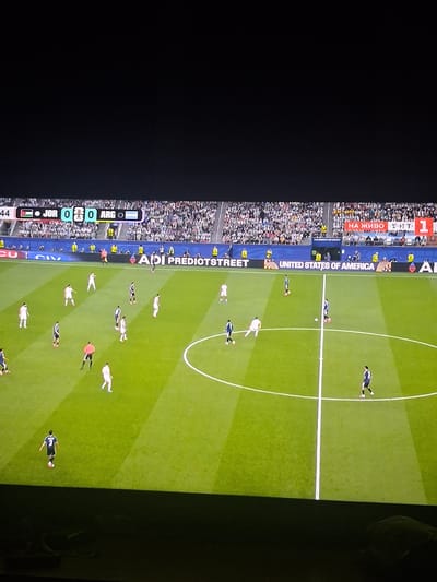 The image captures a soccer match in progress, viewed from a slightly elevated perspective. A digital scoreboard at the top indicates the game is between "JOR" and "ARG" with a score of 0-0. The field is green, marked with white lines, and a circular penalty area is visible. Several players in white and dark blue uniforms are scattered across the field, some actively engaged in play, others observing. Spectators are present in the stands, filling a large portion of the background, many wearing white. Advertising banners are displayed along the edge of the field, with the text "UNITED STATES OF AMERICA" prominently visible. The overall atmosphere appears to be that of a professional sporting event. The city of Krupnik, Bulgaria is not depicted in this image.