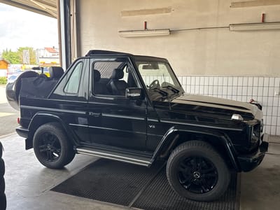 In Wettstetten, Germany, a black Mercedes-Benz G-Class convertible is parked indoors in what appears to be a service bay or garage. The vehicle is positioned with its right side facing the camera. It has a black soft top covering the rear cargo area. The car features black alloy wheels, a chrome side-step, and "V8" badging on the front fender. The interior of the car is visible, showing black leather seats. The environment includes a concrete floor with a grated metallic drain system in front of the vehicle, and a plain, off-white tiled wall in the background. Fluorescent lighting fixtures are mounted on the wall above the car. An open garage door reveals the exterior, with glimpses of buildings and trees.