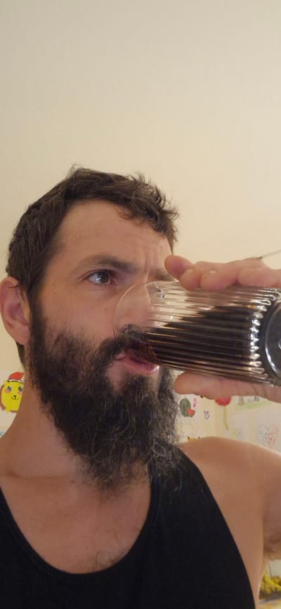 A man with a dark beard and short, wavy hair is drinking from a ribbed glass containing a dark liquid. He is wearing a black tank top. The background appears to be a wall with children's drawings on it, including a yellow creature with a red hat. The image is a close-up, focusing on the man's face and the drink. The location of Wattenwil, Switzerland, is not discernible from the visual information in the image.
