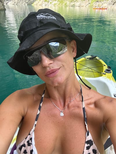 A woman is pictured from the chest up, wearing a black bucket hat with "OUTDOORS" printed in white on the front. She has on large, dark sunglasses that reflect the surrounding scenery, including mountains and a body of water. A thin silver necklace with a heart-shaped pendant rests on her chest. Her bikini top has a leopard print pattern. The woman is in a yellow and white kayak on a bright turquoise body of water, with rocky, mountainous terrain visible in the background. In the distance, another red kayak with two people can be seen on the water. The overall scene suggests an outdoor recreational activity on a lake or fjord, likely in a mountainous region. The question specifies the location as Bezuje, Montenegro.