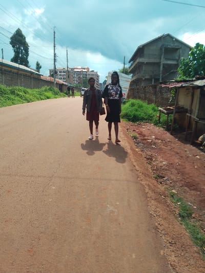 Two young women are walking down a paved road, away from the camera, in Rungiri, Kenya. They are both wearing casual clothing and one is carrying a handbag. There are several buildings visible in the background, including a large apartment building and a two-story house with a fence.  The road is lined with vegetation on both sides, and the ground along the side of the road appears to be dirt. The sky is partly cloudy.