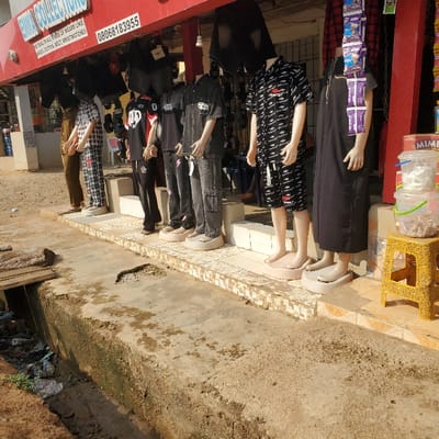 The image depicts a street scene in Eziobodo, Nigeria, specifically in front of a retail shop named "EMMA COLLECTIONS." The shop has a red facade with white lettering and displays contact information, including a phone number "08068183955" and a description: "DEALERS IN ALL KINDS OF WEARS LIKE LADIES, GENTS, BELT, WRISTWATCHES."
Six headless mannequins are positioned along a tiled sidewalk directly in front of the shop, displaying various clothing items. From left to right: one mannequin wears brown trousers (partially visible); another wears black and white checkered pajamas; a third wears a black t-shirt with a white "20" logo and black trousers; a fourth displays a dark short-sleeved shirt over a black t-shirt with dark cargo pants; a fifth wears a black short-sleeved button-up shirt with white logos and matching shorts; and the sixth wears a long black dress. Items, possibly more clothing, hang from the shop's awning. A string of purple-packaged snacks is suspended near the rightmost mannequin.
Adjacent to the sidewalk is a concrete drainage ditch filled with refuse, including plastic bottles, bags, and muddy water. A wooden plank bridges a section of this ditch on the left side. Beyond the ditch is an unpaved, dusty ground area. On the far right, a yellow plastic stool holds stacked clear plastic containers, the bottom one containing what appears to be snacks. The scene is illuminated by bright daylight. No human individuals are visible.