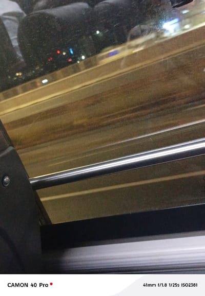 The image depicts an interior view from a moving vehicle, likely a bus or train. The foreground shows a metal handrail and part of a seat. Through the window, blurred city lights and streaks suggest motion and nighttime. Reflections of people are visible in the glass. The camera settings (41mm f/1.8 1/25s ISO2381) indicate low-light conditions and a need for a faster shutter speed to capture movement.