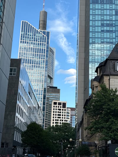 The image depicts a street view in Frankfurt, Germany, characterized by a dense collection of modern skyscrapers and a few older buildings. In the foreground, a dark grey building with horizontal windows lines the left side of the frame. Adjacent to it, a taller building with a white facade and numerous vertical windows dominates the center, featuring a distinctive angled top. Further to the right, a large skyscraper with a glass facade reflecting the blue sky with scattered clouds rises. To the right of the glass skyscraper, an older stone building with a dark, pitched roof and dormer windows is visible. Dense green trees line the street in the mid-ground, obscuring the lower parts of the buildings. The sky is a clear blue with wispy white clouds. In the far distance, several other skyscrapers are visible, suggesting a central business district. The overall impression is one of urban density and architectural contrast, with a mix of contemporary high-rises and historical structures.