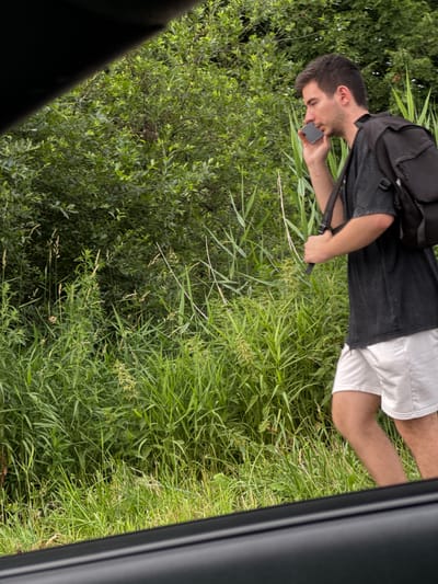 The image shows a young man in a rural outdoor setting, likely in Novoe Devyatkino, Russia. He is wearing a dark t-shirt, white shorts, and a black backpack. He is holding a dark-colored cell phone to his ear and appears to be engaged in a conversation. The man is standing in a grassy area with dense green foliage and tall reeds surrounding him. A dark object, possibly a car door or window frame, is visible in the foreground on the right side of the image, suggesting the photo was taken from inside a vehicle. The lighting suggests it is daytime, and the abundance of green vegetation indicates a mild or warm season. The scene appears calm and natural. There is no visible text in the image.