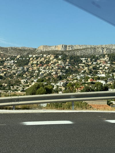 This is a photograph of a densely populated hillside covered in houses, set against a backdrop of rugged mountains. The houses, varying in color from white to yellow and pink, are clustered together, interspersed with trees and vegetation. A highway guardrail runs across the foreground, separating the road from the hillside. The sky is clear and blue. The image appears to be taken from a moving vehicle. Based on the distinct mountainous terrain and the architectural style of the houses, this scene is likely located in Calp, Spain, possibly overlooking the town from a vantage point on the coastal road.