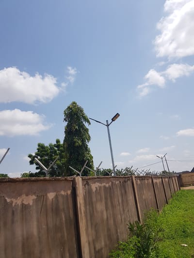 This image captures an outdoor scene with a clear blue sky dotted with white, fluffy clouds, suggesting fair weather during the daytime. The foreground is dominated by a weathered, concrete wall that runs horizontally across the frame, appearing to demarcate a property boundary. Above the wall, a row of sharp, metal anti-climb spikes is visible, indicating a security measure.

To the left of the wall, a tall, slender tree with lush green foliage is prominently featured, its branches swaying, possibly due to a gentle breeze. A street lamp with a distinctive yellow light fixture stands tall next to the tree, mounted on a metal pole. In the background, more trees and greenery are discernible, interspersed with what appear to be utility poles and wires, hinting at an urban or semi-urban environment. The overall impression is one of a typical day in Palade, Nigeria, with a focus on the architectural and natural elements of the setting. There is no visible text in the image.