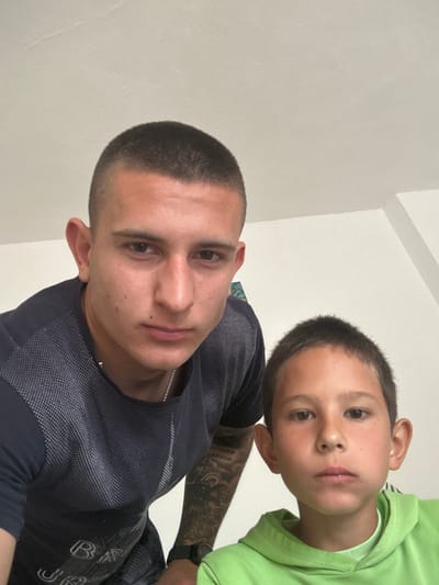 A man and a boy are posing for a close-up photograph, likely a selfie. The man has short, dark hair and is wearing a dark gray t-shirt with a textured pattern. Tattoos are visible on his left arm. The boy is positioned above the man and is wearing a bright green shirt. The background appears to be a plain white wall. The location is Kalchevo, Bulgaria, but there are no features in the image to confirm this.