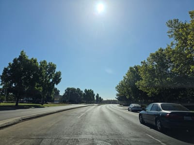 This is a daytime, wide-angle, eye-level shot of a street in Elk Grove, United States of America. The sun is high and bright in the clear blue sky, causing lens flare. The road ahead curves to the right, with a median strip and trees lining both sides. There are at least three cars visible on the road; a dark sedan in the foreground with its license plate reading "LAH279", a lighter colored sedan further ahead, and another lighter colored vehicle in the distance. On the left side of the road, there are several trees and a building with a sign that reads "ROSS" among other retail stores. The road appears to be well-maintained with visible lane markings and a curb. The environment suggests a commercial or suburban area.