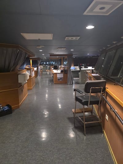 The image depicts the interior of a ship's bridge, likely a vessel docked in Brindisi, Italy, given the context. The scene is dimly lit, with overhead lights casting reflections on the polished gray floor. The bridge is furnished with various consoles, equipment, and furniture. In the foreground, a black captain's chair with chrome legs and a wooden step is positioned near a large windowed section of the bridge, which is partially obscured by reflections. To the left, wooden cabinets and a white air conditioning unit are visible, along with draped gray curtains. The background reveals more consoles and workstations, with glimpses of equipment and papers on desks. The overall environment suggests a functional workspace on a ship, possibly during off-duty hours or while docked.