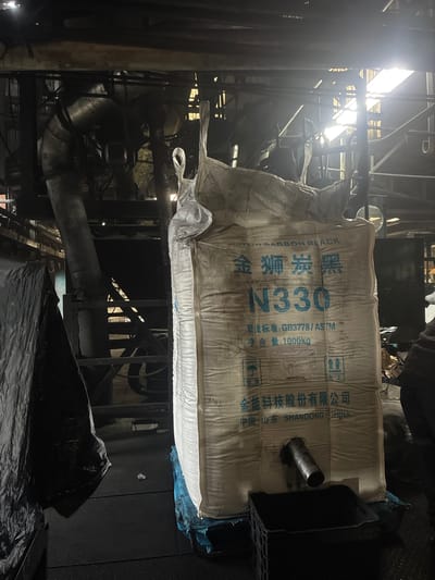 The image shows a large white industrial bag, likely containing carbon black, a powdery substance used in manufacturing tires and other rubber products. The bag is labeled with Chinese characters and the designation "N330," along with "1000kg" indicating its weight. A metal spout protrudes from the bottom of the bag, suggesting it is being discharged into a black plastic container or hopper below. The setting appears to be an industrial facility, with pipes, machinery, and lighting fixtures visible in the background. The overall impression is one of an active production or processing environment. The image does not provide any information to suggest the location is Cuautitlán Izcalli, Mexico.