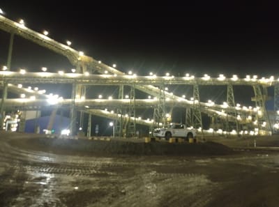 The image depicts an outdoor industrial facility at night. Prominent structures include elevated conveyor belts, supported by a network of green metal scaffolding. Numerous bright lights illuminate the conveyors and surrounding area. In the foreground, a white pickup truck with a light bar on its roof is parked on a mound of dirt. The ground is uneven and appears to be muddy, with tire tracks visible. A large blue structure is visible in the background. The scene is set against a dark night sky. The location is identified as an industrial site within Қопа ауылдық округі, Kazakhstan.