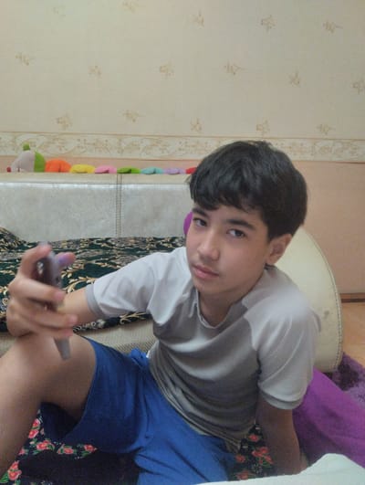 A young male is sitting indoors. He has short dark hair and is wearing a gray and white shirt and blue shorts. He is holding a gray object in his right hand, possibly a phone. Behind him is a bed with a patterned blanket and a white headboard. The wall is decorated with patterned wallpaper. The location cannot be determined as Khujand, Tajikistan.