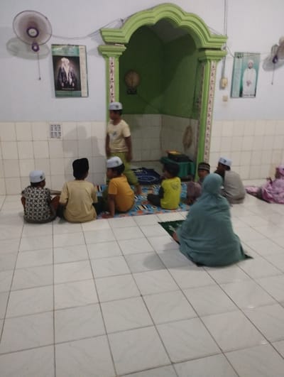 The image depicts an indoor scene within what appears to be a prayer room or small mosque in Kecamatan Arjasa, Indonesia. Several children and a few adults are present. Most of the children wear white peci (Muslim prayer caps), and an adult woman is seated on a green prayer mat, wearing a light blue hijab. Another adult in a grey shirt and white peci is seated further back.
The group is gathered on a light-colored tiled floor, with several children sitting on patterned prayer mats. A taller boy stands near a prominent decorative green arched alcove, which resembles a mihrab, with a few younger children seated on mats in front of him. This setup suggests a religious lesson, Quranic study, or a quiet gathering for prayer.
The room features white walls, with a lower section covered in white tiled wainscoting. Two wall-mounted fans are visible, one on the left and another partially on the right side of the room. Two framed pictures or posters, each displaying a bearded man dressed in traditional attire, hang on the walls. Inside the green alcove, a round, possibly decorative or clock-like object can be seen. The overall atmosphere appears calm and focused. No distinct text is clearly legible in the image.