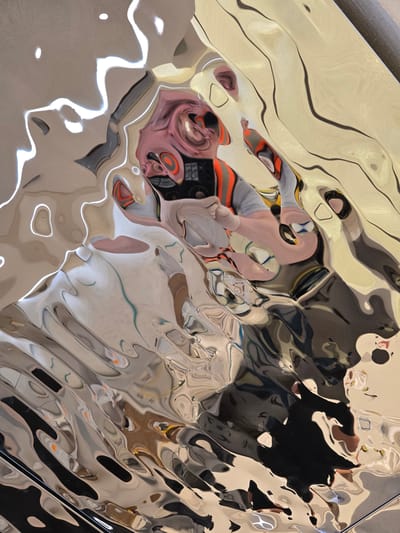 The media file displays an abstract composition formed by reflections on a highly distorted and polished metallic or chrome surface. The surface itself is undulated, creating a warped and fluid effect on all reflected elements.
The primary subject is a person, likely the photographer, captured in the reflection. Their face, visible in the lower-left area, is heavily stretched and distorted, showing pinkish skin tones. Part of a smartphone or camera is reflected, held by hands, indicating the act of photography. The person's clothing reflects as segments of orange and grey fabric.
The surrounding environment reflects as a mosaic of abstract shapes, colors, and varying light intensities. There are smooth, light beige areas interspersed with darker, more angular forms and bright white highlights, suggesting light sources from above or around the scene. The reflections create a sense of depth and movement across the surface.
No specific time of day or weather conditions can be definitively determined from the image, though the lighting appears to be interior or diffuse. There are no discernible architectural features or natural landscapes beyond the abstract reflections. No visible text is legible within the image. Given the location context of Al Jizah, Jordan, the reflective surface could be part of a modern art installation, a decorative architectural element, or a piece of furniture, designed to create visual interest through distortion and reflection. The image is a study in optical illusion and abstract photography.