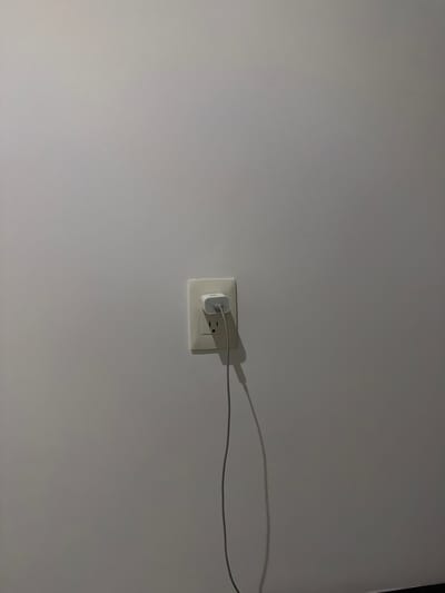 A white wall contains an electrical outlet with a white charger plugged into it. A gray charging cable extends from the charger. The environment appears indoors. There is no verifiable information about the location being in Villahermosa, Mexico. No people, subjects, or actions are visible.