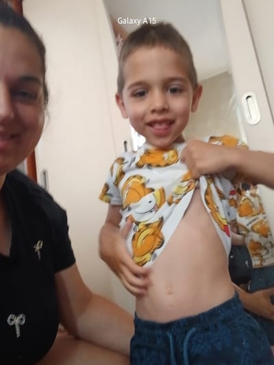 An indoor, vertically oriented close-up photograph captures a playful interaction between an adult and a young child, likely a boy, in Obrochishte, Bulgaria. The image is taken from a low angle, looking upwards at the subjects, which gives them a tilted orientation within the frame.
At the top of the photograph, an adult is partially visible, wearing a black top with small, shiny silver or white decorative elements, possibly bows or star shapes. The adult's face is partially seen on the upper right, looking towards the camera with a gentle expression.
The main subject is a young boy, positioned prominently in the center. He has short, light brown hair and is smiling broadly at the camera. He is wearing a white t-shirt adorned with multiple orange cartoon cat characters, resembling Garfield, and dark blue shorts. The boy is actively lifting his t-shirt with both hands, playfully revealing his bare stomach and belly button.
The background consists of light-colored walls or furniture, possibly cabinets or a door. A silver or white recessed handle is visible on the right side of this background element. The lighting suggests an indoor setting during the day or in a well-lit interior. The visible text "Galaxy A15" is present vertically along the right edge of the image, indicating the camera device used to capture the photo. The overall scene conveys a cheerful and informal atmosphere.
