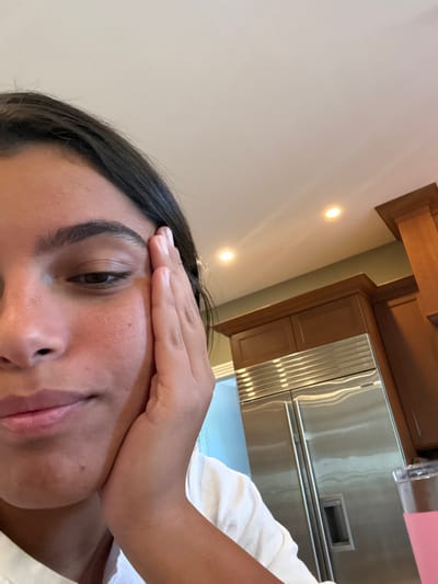 The image depicts an indoor kitchen setting. A single individual, appearing to be a young female with dark hair and medium skin tone, occupies the lower-left portion of the frame. Her left hand is resting against the left side of her face, near her temple and cheek. She is wearing a light-colored top, and her visible eye is cast downwards, indicating a neutral or contemplative expression.
In the background, to the right of the individual, a large stainless steel side-by-side refrigerator is visible, featuring vertical handles and horizontal ventilation grilles. Above and to the right of the refrigerator, light brown wooden kitchen cabinets are mounted on a wall painted light green. The ceiling is white, with two illuminated recessed spotlights visible. In the bottom-right corner, a clear tumbler containing a pink liquid is partially visible. A bright, undefined light source or reflection appears to the left of the refrigerator. The specific location within Princeton, United States, cannot be determined from the visual information presented.