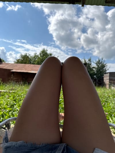The image depicts a pair of human legs from the knees down, positioned against a backdrop of a rural landscape under a cloudy blue sky. The legs are bare and appear to be tanned. The person is wearing denim shorts with frayed edges. In the background, there is lush green vegetation, including grass and wildflowers, with trees and rustic wooden structures, possibly barns or outbuildings. A metal chair frame is visible at the bottom left of the image. The scene suggests a casual outdoor setting, likely in a village or countryside area. The location of Малдыкасы, Russia, is not explicitly visible or indicated in the image itself, but the overall environment is consistent with a rural Russian setting.
